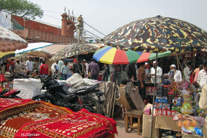 Eine belebte Szenerie mit Schirmen, Fahrzeugen, Matten, Kisten, einem Hocker, Menschen und diversen anderen Gegenständen, mit einer Moschee, Mikrofonen, einem Strommast und Kabeln im Hintergrund.