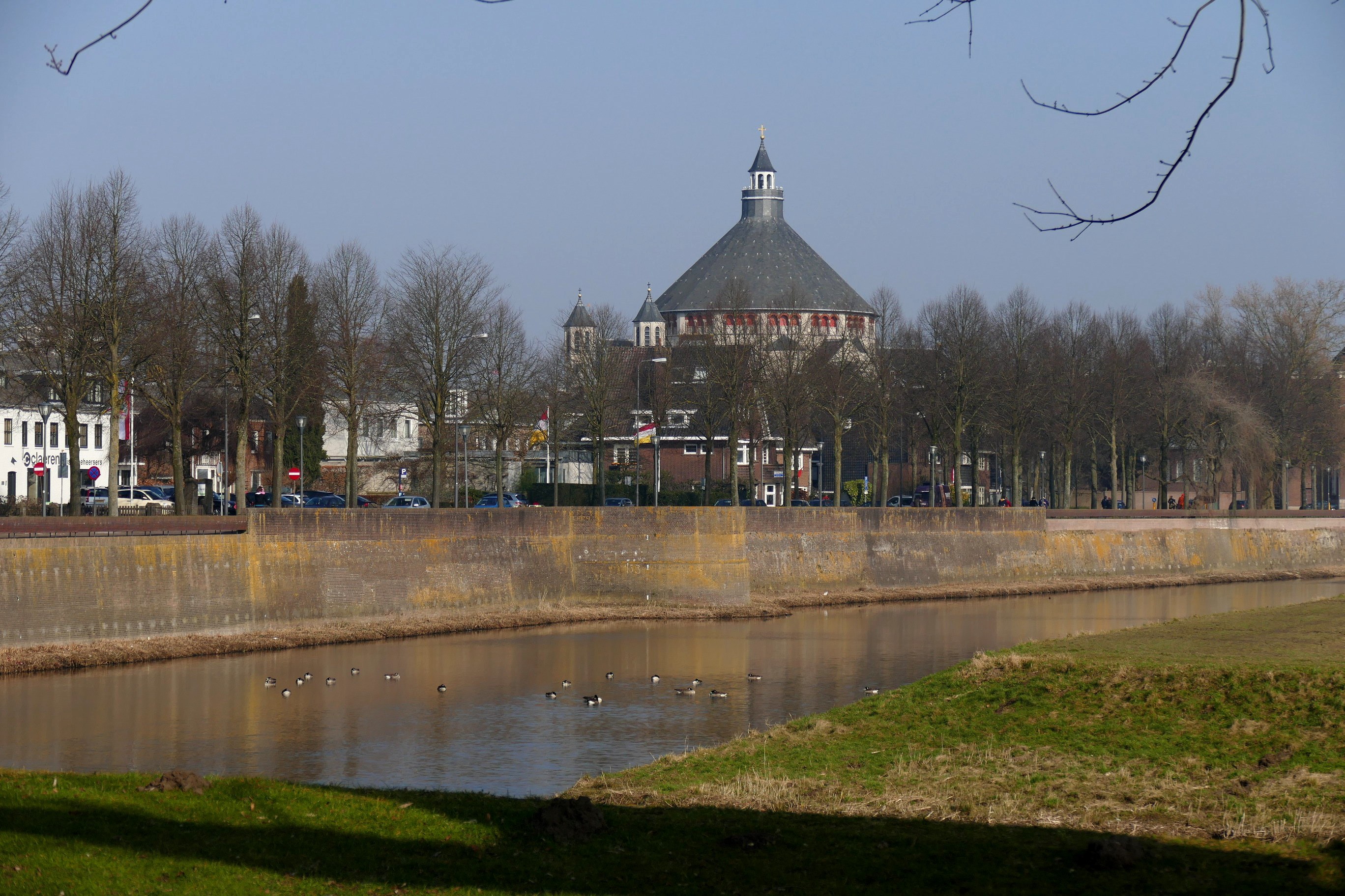 Ein Fluss mit schwimmenden Enten, umgeben von grünem Gras und Bäumen, mit Gebäuden, Fahrzeugen, Pfählen, Flaggen und einem klaren blauen Himmel im Hintergrund.