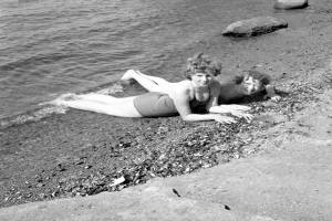 Zwei Frauen in Badesachen liegen auf einem Strand umgeben von Steinen und glitzerndem Wasser, dargestellt in Schwarz-Weiß.