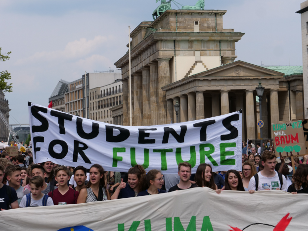 Eine Gruppe von Schülern marschiert in Berlin mit einem bunt bemalten Banner, auf dem "Students for Future" steht, vor einem Hintergrund aus Gebäuden, Bäumen und Himmel.