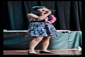 Eine Frau in einem bunten Kleid singt auf der Bühne in ein Mikrofon, mit einem Tisch und Papieren im Hintergrund hinter einem Vorhang.