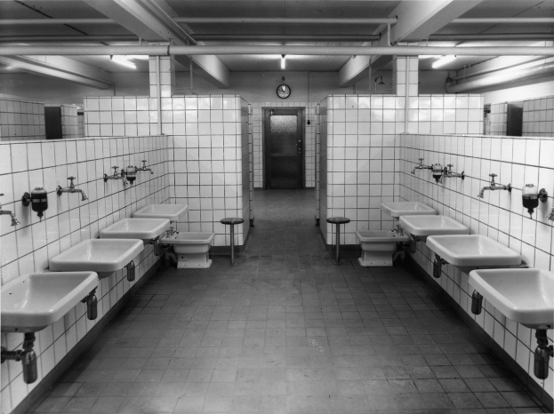 Schwarz-weiß-Foto einer öffentlichen Toilette mit Waschbecken, Wasserhähnen, Hockern, einer Wanduhr und Deckenleuchten.