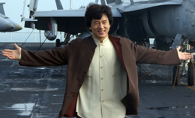 Jackie Chan steht auf dem Deck eines Flugzeugträgers mit ausgebreiteten Armen in einem weißen und braunen Kleid, während im Hintergrund Kampfflugzeuge und ein klarer blauer Himmel zu sehen sind.