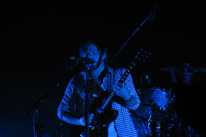 Ein Mann in der Mitte des Bildes spielt Gitarre und singt in ein Mikrofon, während hinter ihm Schlagzeug in einer dunklen Konzerteinrichtung zu sehen ist.