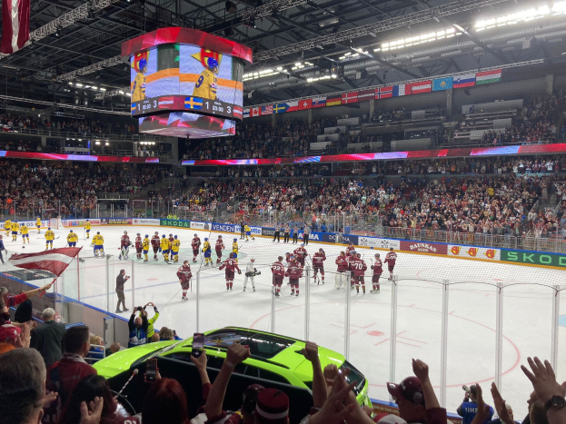 Hockey game in a large arena with spectators, rink fencing, banners, ceiling lights and screen, and flags on the ceiling and held by attendees, with a parked car visible outside the rink.