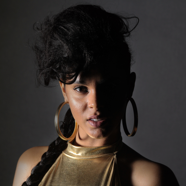 Eine Frau in einem goldenen Kleid und großen Ohrringen steht selbstbewusst vor einer Wand, ihr glatter Bob-Haarschnitt und ihr natürliches Make-up unterstreichen ihren entschlossenen Gesichtsausdruck, während sie direkt in die Kamera blickt.