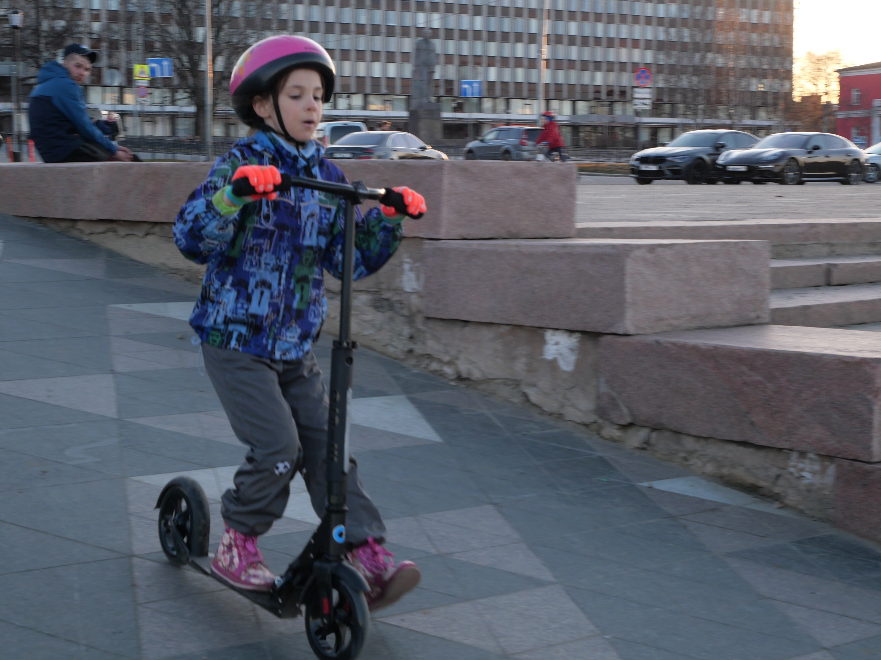 Ein junger Junge in Helm und Handschuhen fährt auf einem Roller eine Straße entlang, mit Stufen, Fahrzeugen, Menschen, Bäumen, Pfählen, Brettern, Gebäuden und einem klaren blauen Himmel im Hintergrund.