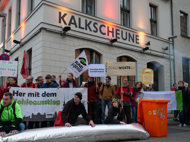 Gruppe von Menschen mit Schildern und Plakaten vor einem Gebäude während einer Demonstration in Deutschland, mit zwei Personen im Vordergrund und einem Müllcontainer rechts.