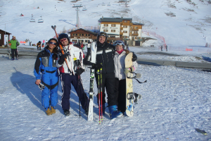Vier Personen posieren für ein Foto mit Schlittschuhausrüstung und Werkzeugen im Hintergrund, mit Gebäuden und anderen Menschen vor einer schneebedeckten Berglandschaft.