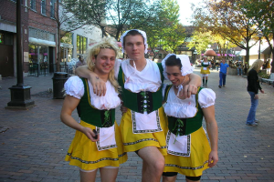 Drei Männer in weißen und gelben Kostümen stehen lachend vor einer belebten Straßenszene mit Menschen, Gebäuden, Bäumen, einer Bank, einem Müllcontainer und einem Geländer.