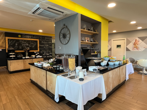 Ein Restaurantinnenraum mit Tischen und Stühlen, Lebensmitteln, einer Tafel mit Text, einer Wanduhr, Deckenleuchten, einer Klimaanlage und einer Tür im Hintergrund, Located im Hôtel de Ville in Paris.