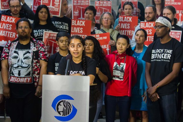 Eine Gruppe von Menschen mit "Black Lives Matter"-Plakaten, eine Frau spricht vor einer Menge an einem Rednerpult und eine Wand im Hintergrund.