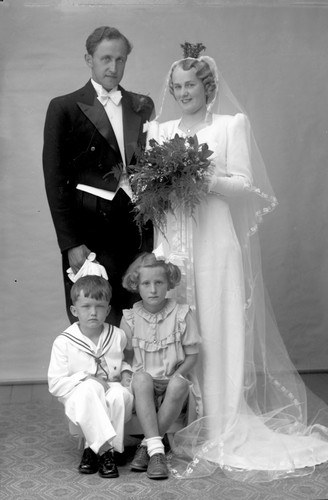 Schwarzes und weißes Foto einer Familie mit einem Hund, die vor einem Haus posieren, einschließlich eines Mannes, einer Frau und mehrerer Kinder.