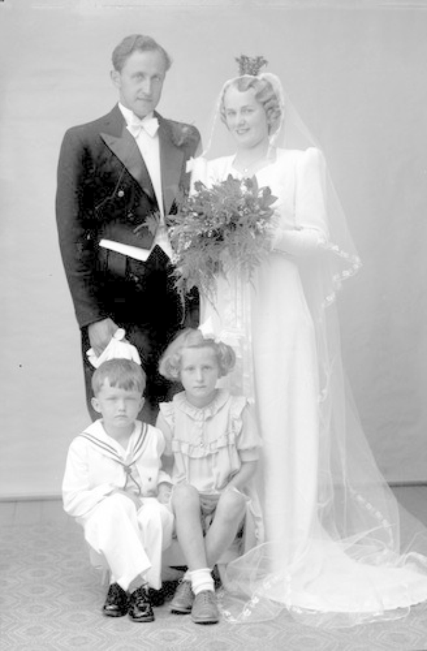 Schwarzes und weißes Foto einer Familie mit einem Hund, die vor einem Haus posieren, einschließlich eines Mannes, einer Frau und mehrerer Kinder.