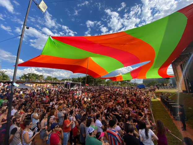 Eine gro%C3%9Fe Menge steht vor einer Regenbogenflagge bei einer Festivit%C3%A4t, mit einem Geb%C3%A4ude, einer B%C3%BChne und B%C3%A4umen im Hintergrund.