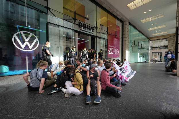 Menschen sitzen vor einer Volkswagen-Niederlassung auf dem Boden und halten ein Transparent.