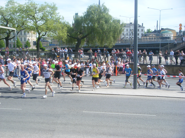 Eine Gruppe von Menschen, die bei einem Marathon auf einer Straße laufen, umgeben von einem Metallzaun, einem Torpfosten, einer Schleife an einem Pfahl, Zuschauern auf dem Gehweg, Metallabsperrungen, Pfählen, Straßenpfählen, Schildern, einer Brücke, Gebäuden mit Fenstern, Bäumen und einem bewölkten Himmel.