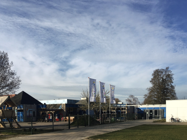 Gro├če Geb├Ąude mit einem grasigen Bereich vorne, umgeben von einem Metallzaun, B├Ąumen, Bannern, einem geparkten Fahrrad, n├Ąheren H├Ąusern und einem bew├Âlktem Himmel, als das neue Kinderspielgel├Ąnde an der Schule identifiziert.