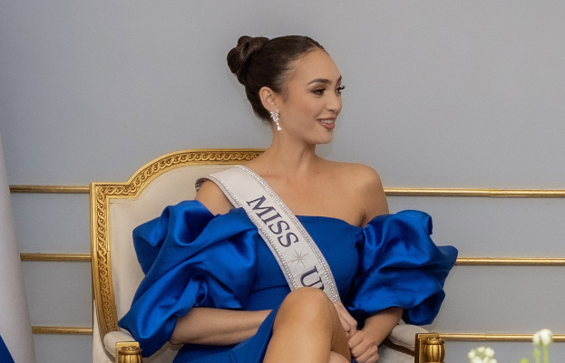 Eine Frau in einem blauen Kleid trägt ein 'Miss Universe 2019'-Band und sitzt lächelnd auf einem Stuhl umgeben von Blumen mit einer Wand im Hintergrund.