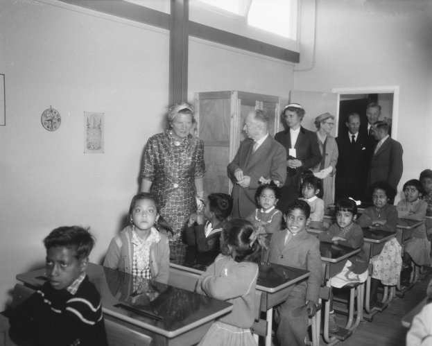 Schwarzes und weißes Foto einer Klasse mit Kindern auf Bänken sitzend und stehend, eine Frau in der Mitte, eine Uhr an der Rückwand, Papiere auf einem Schrank, Tür und Decke, sowie Deckenleuchten.