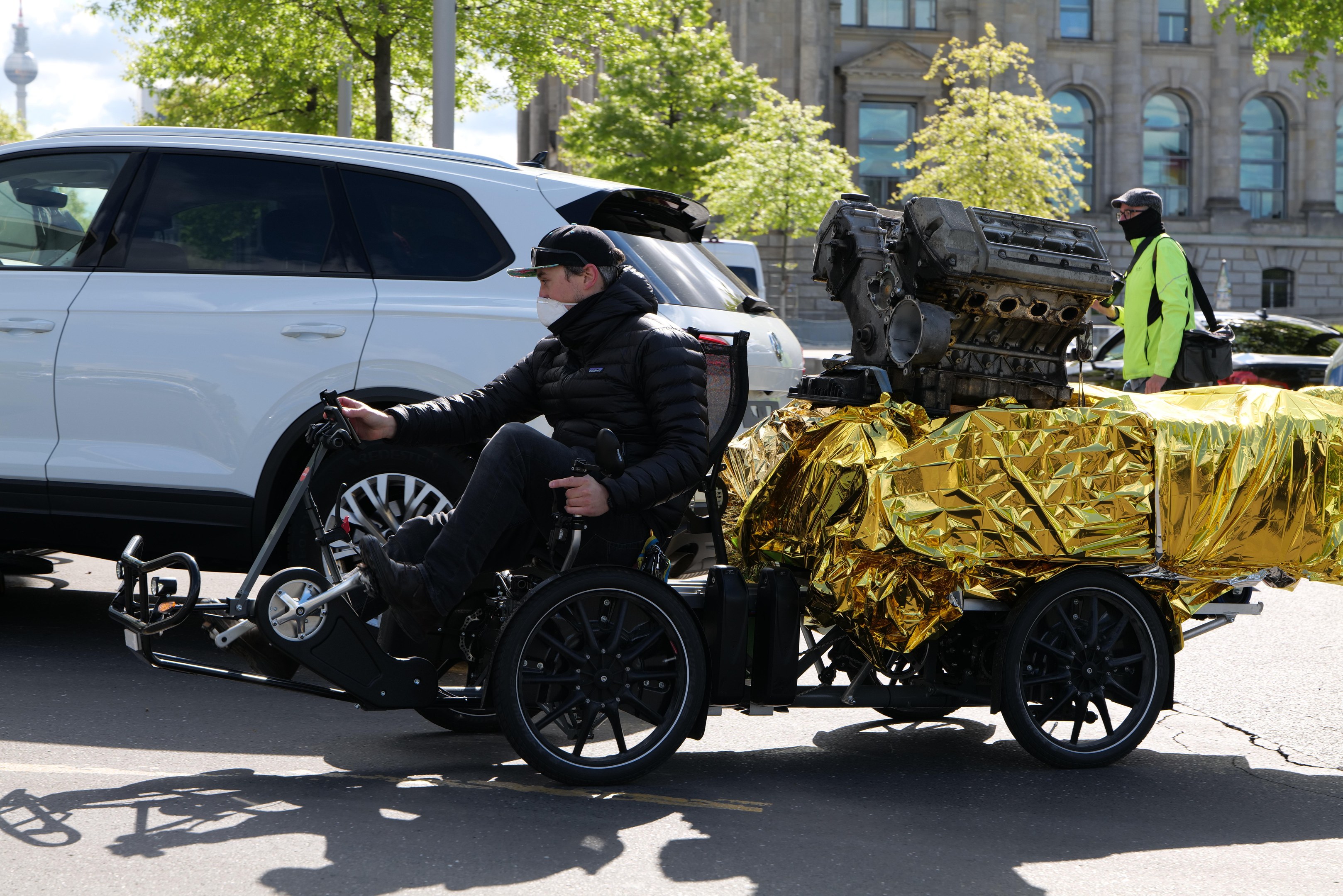 Ein Mann im Rollstuhl mit einem großen Motor daran, umgeben von Fahrzeugen auf einer Straße mit Bäumen, Gebäuden und einem klaren blauen Himmel im Hintergrund, trägt eine schwarze Jacke und eine Mütze und hält ein Objekt.