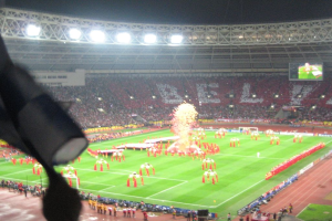 Ein Stadion mit Flutlicht, Eisengittern, Zuschauern, stehenden Personen, Werbetafeln, Bildschirmen, Überwachungskameras und dekorativen Fahnen.