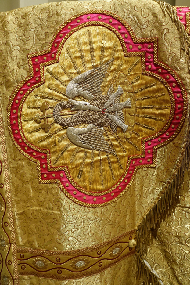Nahaufnahme eines Priestergewandes mit aufwendiger Gold- und Rotstickerei vor einer Wand im Hintergrund.