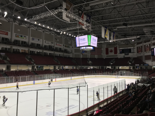 Eishockeyspiel in einer großen Arena mit einem zentralen Netz, Zuschauern auf Stühlen, Deckenbeleuchtung und Trägern, Bannern und Fahnen; die Arena ist als St. Louis Blues Arena identifiziert.