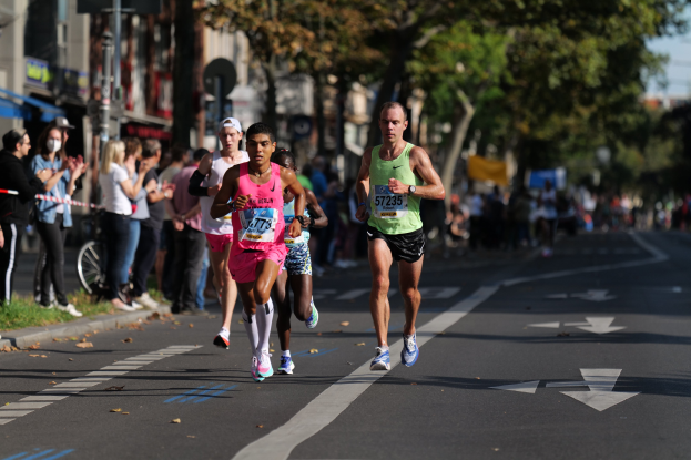 Gruppe von Menschen, die in einem Stadtmarathon laufen, mit Zuschauern auf der linken Seite, unscharfen städtischen Hintergrund mit Bäumen, Gebäuden und einem Fahrrad.