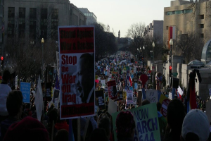 Eine Gruppe von Menschen demonstriert auf einer Straße und hält Plakate in den Händen, im Hintergrund sind Gebäude, Bäume, Laternenpfähle und der Himmel zu sehen.