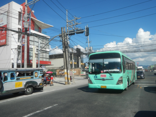 Ein grüner Bus fährt auf einer Straße mit hohen Gebäuden, Fußgängern auf dem Gehweg mit Regenschirmen und Stromleitungen oben.