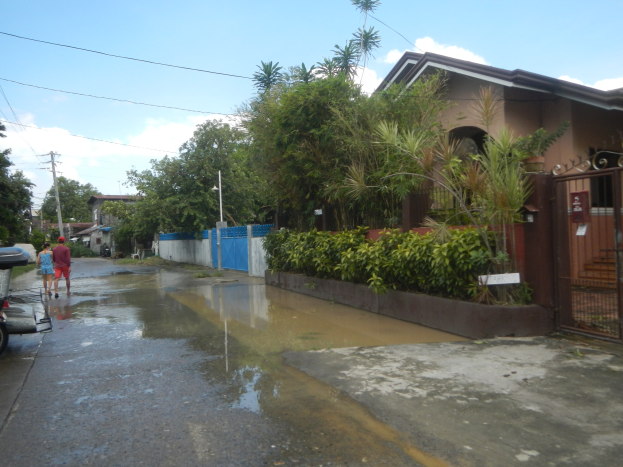 Eine überflutete Straße mit einem geparkten Motorrad am Straßenrand, Menschen, die gehen, Gebäude, Bäume, Pflanzen, Pfosten, Drähte, ein Tor im Hintergrund und ein bewölkter Himmel mit einem Wasserzeichen.