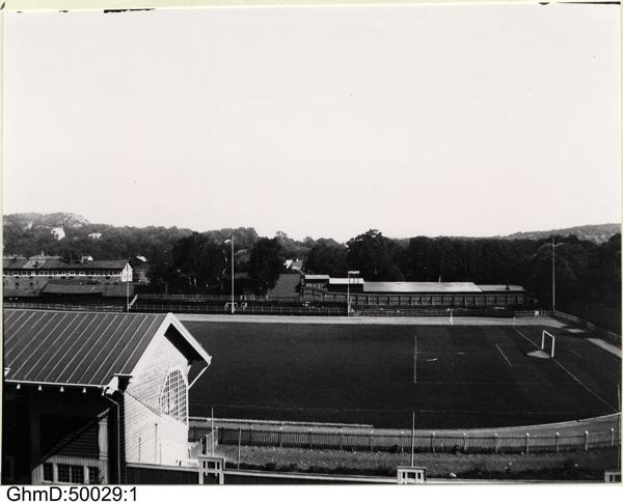 Schwarzes und weißes Foto eines Fußballfeldes mit einem Schuppen links, einem umgebenden Zaun, zentralen Pfosten und Bäumen mit Himmel im Hintergrund, beschriftet mit "Gillingham Football Club, 1960er Jahre" unten.