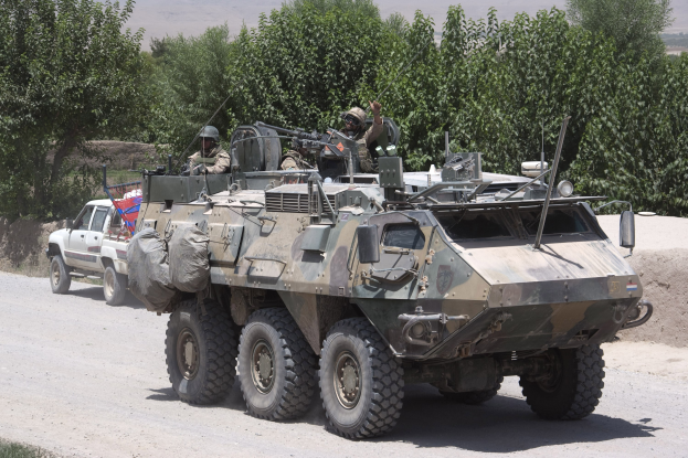 Ein gepanzertes Mannschaftsfahrzeug und ein Lastwagen fahren auf einer Schotterstraße, mit bewaffneten Personen in Helmen in der Nähe, vor einem Hintergrund aus Bäumen, Hügeln und einem klaren Himmel.
