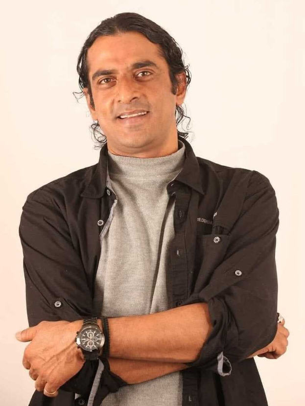 Ein Mann in einem schwarzen und aschfarbenen Kleid mit überkreuzten Armen, der eine Uhr trägt und vor einem weißen Hintergrund steht, der in Gedanken versunken scheint.