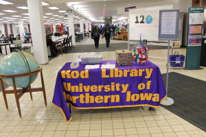 Eine Ausstellung mit einem Tisch, auf dem verschiedene Landesflaggen, eine Box, Papiere und eine Fahne sowie links daneben eine Weltkugel, rechts daneben eine Anschlagtafel mit einigen Menschen in der Nähe und mehrere Tische und Stühle in der Ferne zu sehen sind.