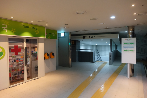 Der Eingang einer Apotheke mit einem "Apotheke"-Schild auf der linken Seite, einer Tafel mit Text auf der rechten Seite und beleuchteten Lampen auf dem Dach.