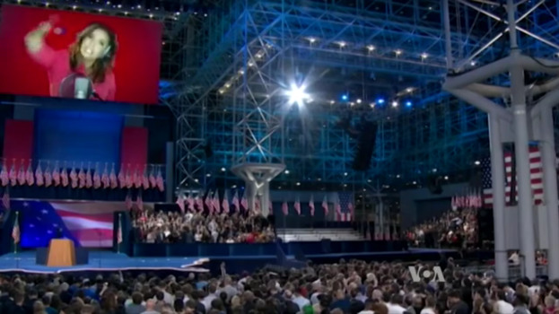 Eine große Menschenmenge vor einer Bühne mit einem Podium, einem Mikrofon, Flaggen, einem Bildschirm mit einer Frau und Lichtausrüstung, wahrscheinlich bei einer Präsidenten-Debatte.