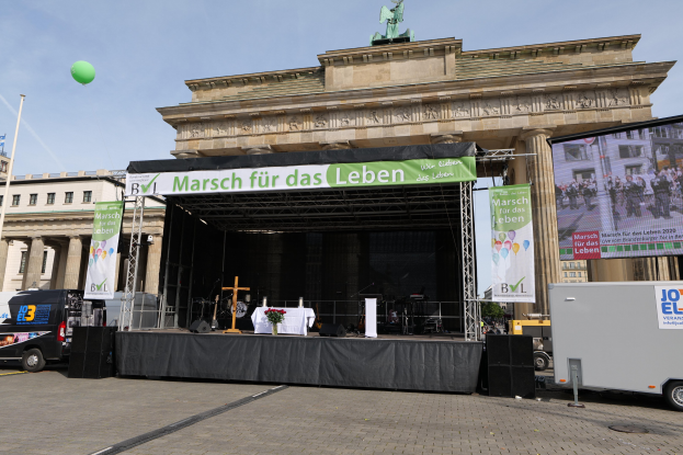 Eine Bühne vor dem Brandenburger Tor in Berlin, Deutschland, mit einem Tisch mit einer Vase, einem Kreuzsymbol, Transparenten, Lautsprechern und anderen Gegenständen, sowie Fahrzeugen auf der Straße und Gebäuden im Hintergrund.