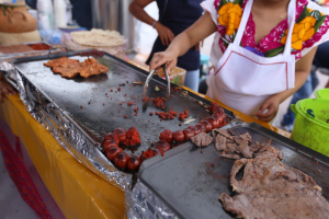 Eine Frau in einer Schürze grillt Essen auf einem Markt, mit einem Tisch voller Lebensmittel, einem Eimer und anderen Gegenständen in der Nähe und einigen Menschen im Hintergrund.