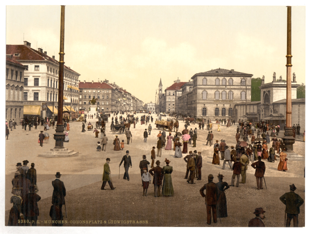 Schwarzes und weißes Foto einer belebten Münchner Straße in den frühen 1900er Jahren mit Passanten, einem Pferdewagen, Gebäuden, Bäumen und einem sichtbaren Himmel.