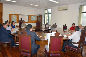 Eine Gruppe von Menschen sitzt an einem Tisch mit Papieren und Gläsern, einem Klimagerät, einer Anschlagtafel und Karten an der Wand im Hintergrund.