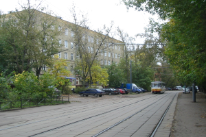 Eine Straßenbahn fährt auf einer von parkenden Autos gesäumten Straße, umgeben von Bäumen und einem Geländer, mit Gebäuden und einem klaren Himmel im Hintergrund.