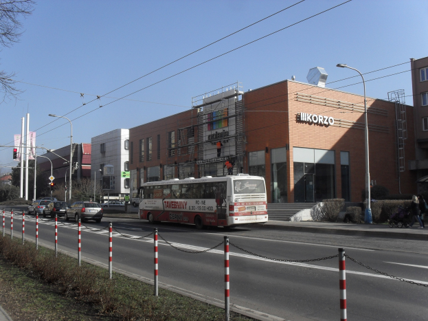 Ein Bus fährt eine Straße mit hohen Gebäuden, Straßenlaternen, Strommasten, Stromkabeln, Kraftfahrzeugen, Barrieren, Fußgängern, Büschen, Bäumen und einem klaren blauen Himmel entlang.