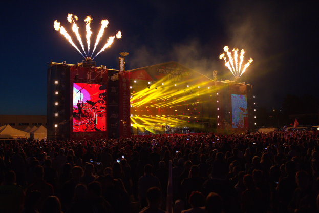 Eine große Menschenmenge bei einem Musikfestival schaut auf der Bühne mit bunten Fahnen und Feuerwerk am dunklen Himmel.