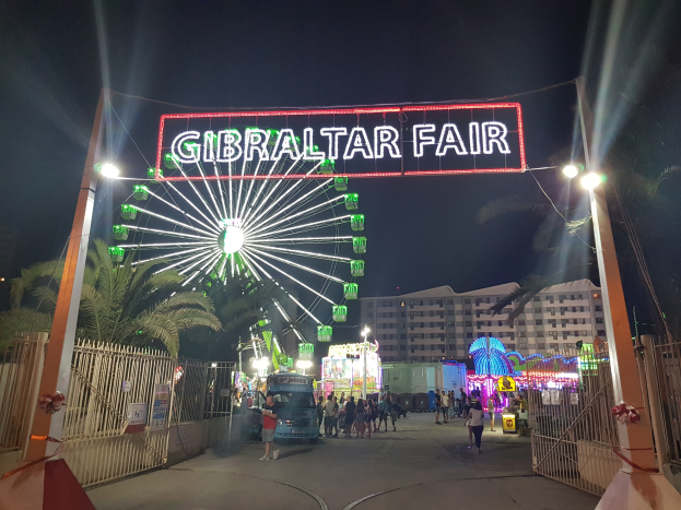 Der Eingang zur Gibraltar-Messe bei Nacht mit einem hell erleuchteten Riesenrad und einer leuchtenden Namensplakette, umgeben von Menschenmassen, Bäumen und Gebäuden im Hintergrund unter einem dunklen Himmel.