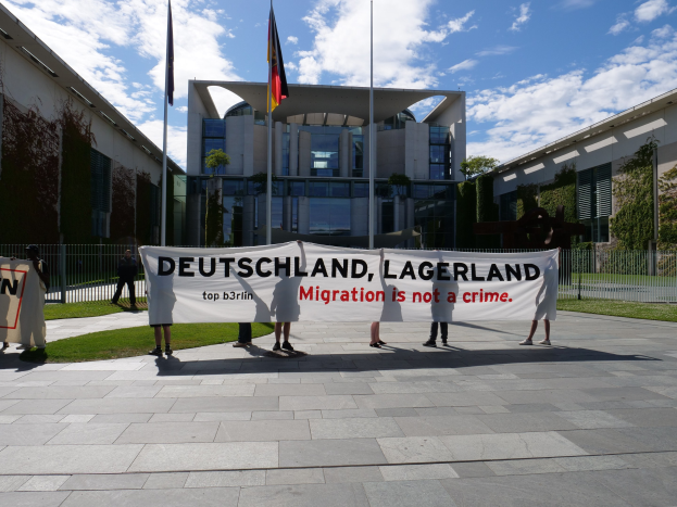 Eine Gruppe von Menschen mit einem Transparent, auf dem "Deutschland, Lagerland Migration ist kein Verbrechen" steht, vor einem Gebäude mit Fenstern, umgeben von Gras, einem Metallzaun, Pflanzen, Bäumen und Fahnen mit Ständen, unter einem bewölktem Himmel.