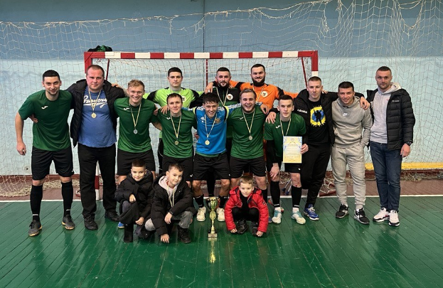 Eine Gruppe von Männern in Medaillen, einer hält einen Pokal, steht auf einem Futsal-Platz mit einem Tor und einer Wand im Hintergrund, feiert ihren Futsal-Weltmeister-Titel.