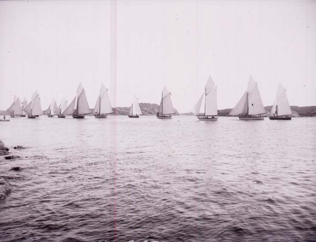 Schwarzes und weißes Foto von Segelbooten auf dem Wasser mit Bergen im Hintergrund und einem klaren Himmel, beschriftet als New York Yacht Club.