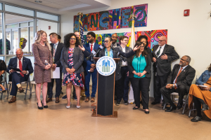 Eine Gruppe von Menschen, die in einem Raum mit einem Podium, einem Mikrofon und einer Tafel mit der Aufschrift "Sancho-Stadtrat tritt dem Stadtrat bei" stehen und sitzen, mit Flaggen, einer Tafel, einem Feuerlöscher und Deckenleuchten an der Rückwand.